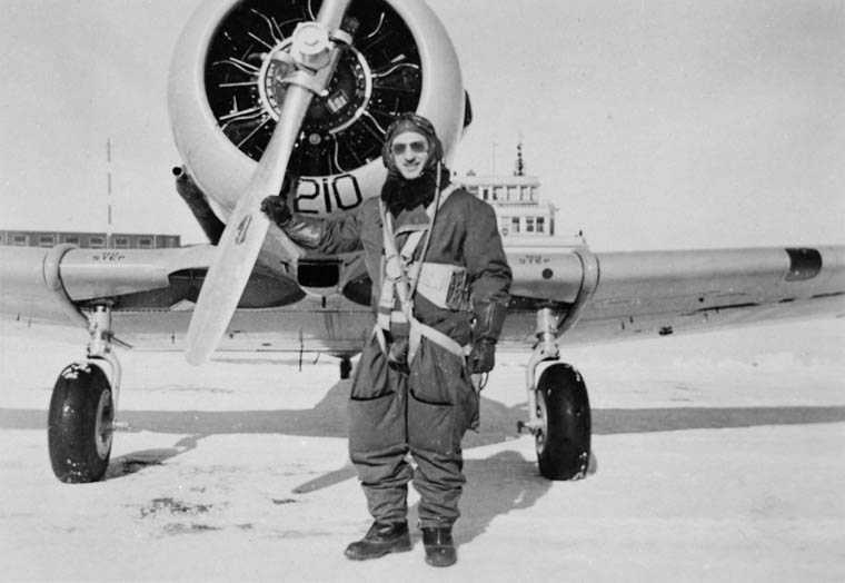 Black and white photograph. A man in heavy pilots gear stands in front of an airplane, leaning one arm on the propeller.